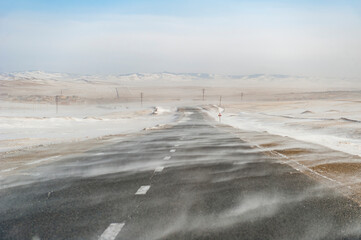 Leere Landstra&szlig;e in der winterlichen Steppe der Mongolei aus Fahrersicht mit Schneeverwehungen und einer Siedlung und Bergen am Horizont