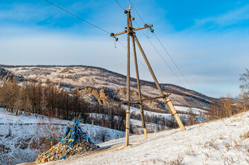 H&ouml;lzerner Strommast mit &Uuml;berlandleitung in menschenleerer, mongolischer Winterlandschaft und einem sog. "Owoo", einem verzierten Steinhaufen mit Gebetsfahnen f&uuml;r religi&ouml;se Zeremonien