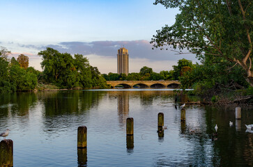 View of a city building over a calm river with trees and a bridge in the background during early evening hours .