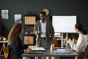 Black man standing and presenting business charts to two young adult women in modern office, colleagues listening and discussing data, digital screen displaying graphs in background