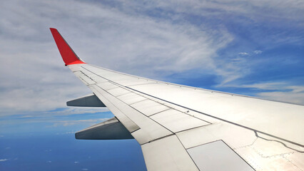 the view from airplane window, showing the wing of an aircraft with distinctive red winglet against a blue sky with white cloud