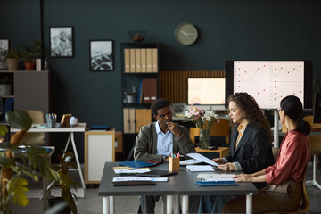 Multiethnic group of young adult professionals discussing business documents at office table, analyzing financial charts on digital screen, collaborating on project in modern workspace