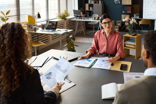 Caucasian woman leading business meeting with multiethnic colleagues discussing financial charts and reports in modern office setting, participants sitting around table - Powered by Adobe