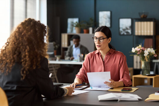 Caucasian woman wearing glasses discussing documents with young adult woman across desk in modern office, male colleague working in background, business meeting scene