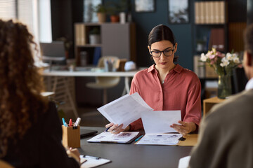 Caucasian young adult woman reviewing documents during business meeting with multiethnic colleagues in modern office, sitting at desk and focusing on paperwork while others listening