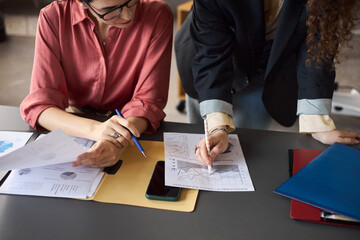 Caucasian young adult women analyzing financial charts and graphs at desk, reviewing printed documents and taking notes with pens during business meeting