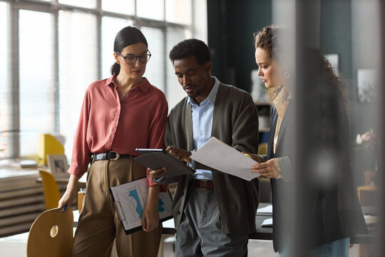 Caucasian women and Black man collaborating in modern office, reviewing documents and digital tablet, discussing project details together