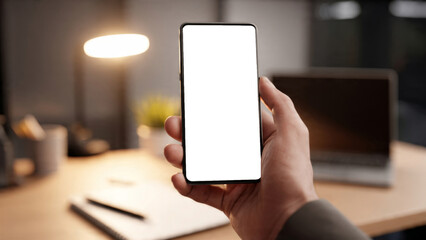 Hand Holding Smartphone with Blank Display in Modern Office Setting with Laptop and Desk Lamp