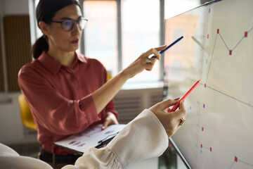 Young adult Caucasian woman explaining data on digital screen while young another woman holding clipboard analyzing graph, both collaborating on business presentation