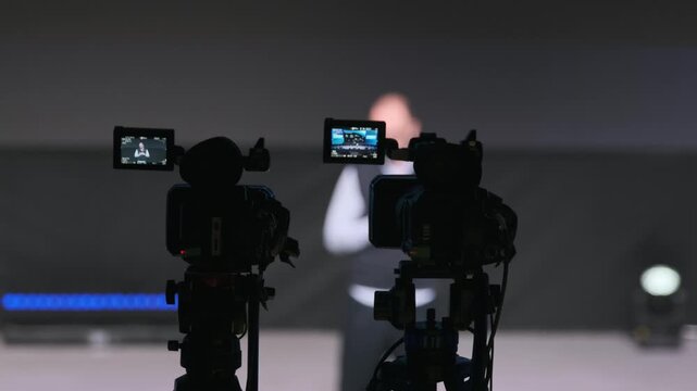 Two cameras on tripods film speaker on stage during studio shoot. Dim background, spotlight, focus on filming process, professional media event, interview style, audience not visible
