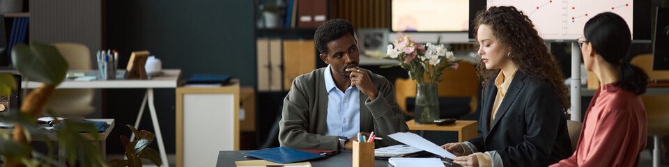 Multiethnic group of professionals discussing business documents at office table, Black man listening attentively while Caucasian woman explaining project details to another woman, header