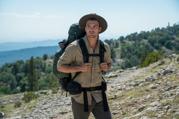 Adventurous male hiker with a large pack and wide brim hat standing on a rocky mountain path, looking at the scenic valley view during a bright sunny day.