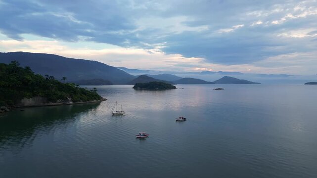 Paraty, Brazil: Aerial drone footage of boat sailing in atlantic ocean in Paraty, an old town in state of Rio de Janeiro, Brazil during cloudy sunset with mountain in the background . Taken with orbit