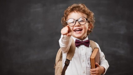 Happy student boy laughing and pointing at the camera. Cheerful child with glasses and a book in front of a chalkboard. Back to school and education concept with copy space
