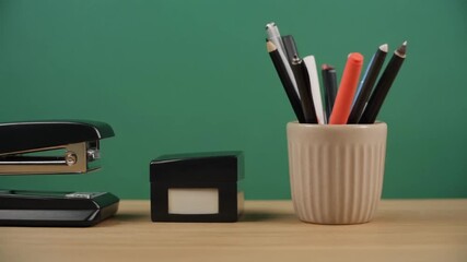 Various office supplies including a stapler, pens, and pencils on a wooden desk with a green background.