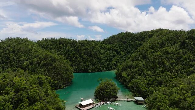 Emerald green lagoon lies between lush tropical hills beneath partly cloudy blue sky. Sugba Blue Lagoon. Siargao, Philippines.