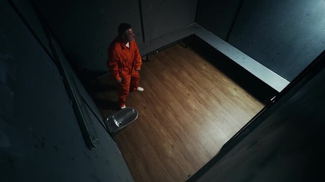 Aggressive male inmate in jumpsuit pacing prison cell under video surveillance