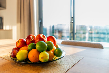 Front View of a Glass Bowl of Mixed Fresh Fruit, Including Apples and Citrus, on a Wooden Table by a Bright Window