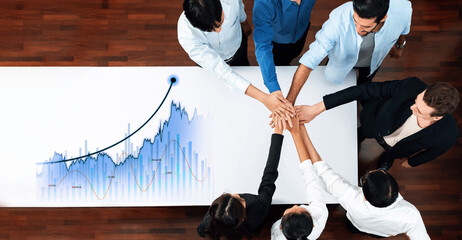A diverse team of professionals gathers around a table, celebrating their financial success with hands united, highlighting the importance of teamwork and collaboration in achieving goals. Scalp