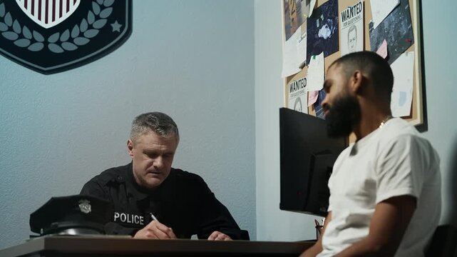 Middle-aged police officer taking notes while questioning male witness in office