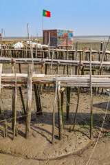 A colorful, rustic wooden fishing shack standing on the traditional stilt pier system of Palafita da Carrasqueira, Portugal, at low tide.