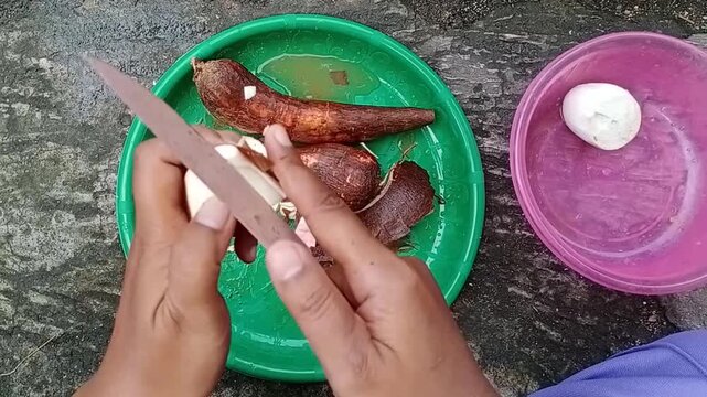 Asian hands cutting and peeling fresh cassava roots with a knife over plastic plate outdoors. Traditional food preparation scene showing raw tubers, natural texture, and simple home cooking process
