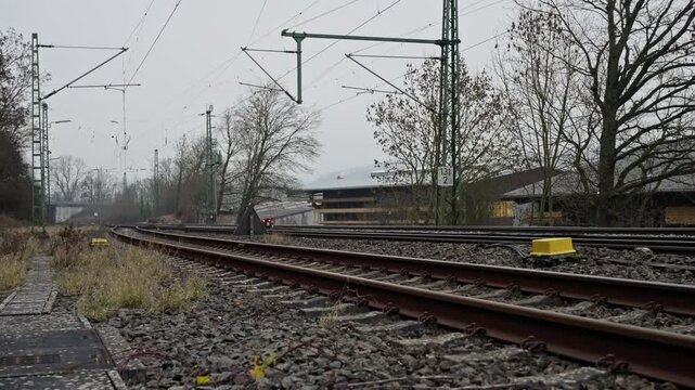 Empty Curved Tracks Under Overcast Sky, Electrified Catenary And Poles Lining Gravel Ballast, Damp Platform And Distant Station Building, Industrial Fringe With Muted Tones And Calm Commuter Absence