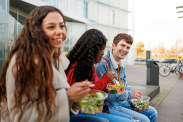 Three multiethnic students enjoying healthy takeaway salads while sitting outdoors