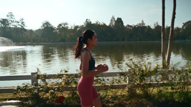 Fit young asian woman running in a park, enjoying her morning workout routine next to a tranquil lake. Healthy lifestyle and fitness concept