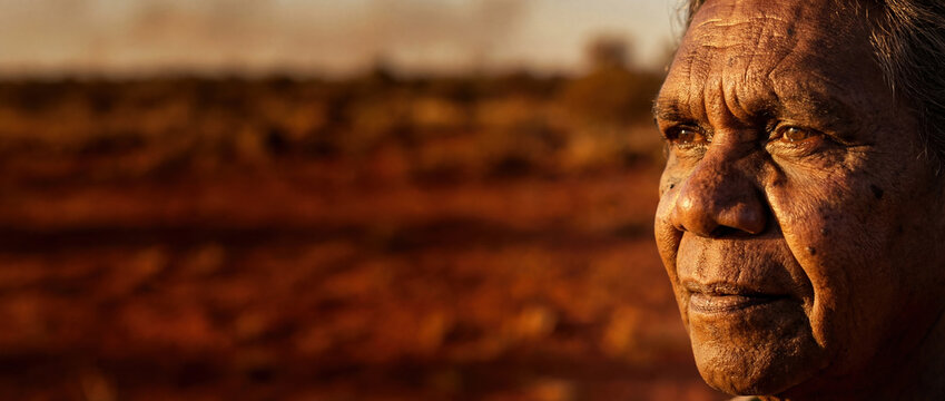 Portrait of an elderly Indigenous Australian woman looking across the outback landscape at sunset. Close-up of an Aboriginal elder's face with red earth background and copy space.
