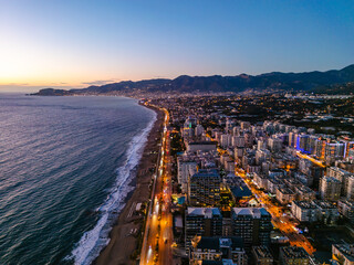 Panoramic Aerial Night View of Alanya Coastline with Illuminated Cityscape and Mountain Range at Twilight