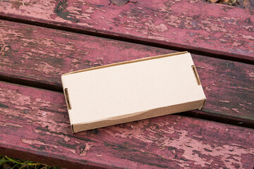 Small blank cardboard box lying on a weathered red wooden bench with peeling paint. Delivery, parcel, packaging, and e-commerce concept with copy space.