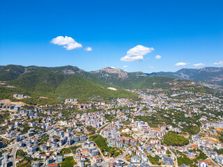 High-Angle Aerial View of a Dense Coastal Residential Area Nestled Against Lush, Tree-Covered Mountains Under a Clear Blue Sky