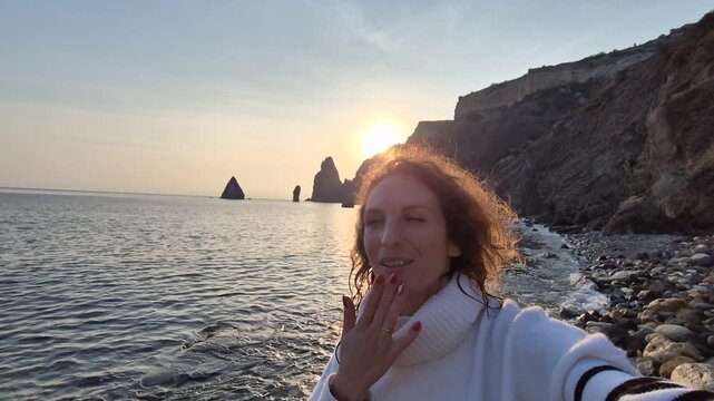 Woman sunset beach joyful travel blogger films selfie video laughing and waving goodbye on a rocky coastline.