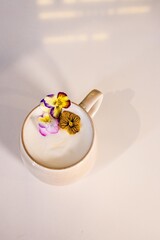 Minimalist coffee cup with creamy milk foam and edible pansy flowers. Soft natural lighting, neutral tones and clean composition create a calm, elegant mood suitable for modern design and lifestyle us