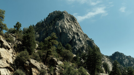 Majestic mountain peak covered in rugged rocks and lush green trees stands tall under a clear blue sky with scattered wisps of clouds on a sunny day in nature.