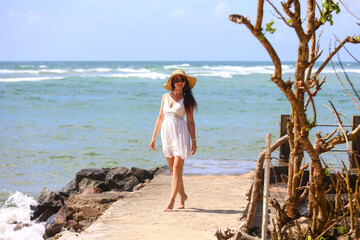 Woman in white dress and hat walking by the ocean on a sunny day