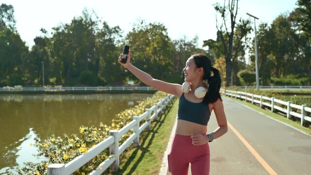 Cheerful Asian runner live streaming or having a video call on her smartphone while jogging. Vlogger creating content for her social media