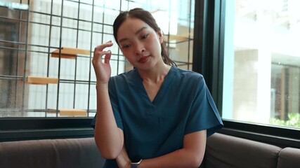 Young female doctor wearing scrubs and sitting in a modern office, feeling tired and stressed during a difficult video conference on her laptop
