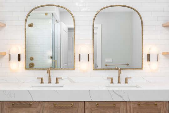 A bathroom detail with gold faucets, light fixtures, and mirrors, a wood cabinet, large marble slab countertop, and a subway tile wall.