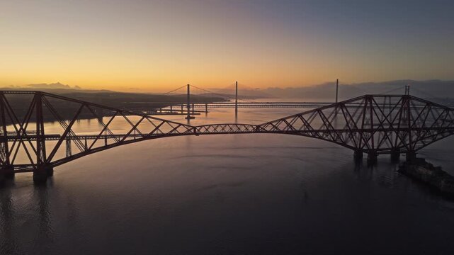 Aerial view of a red steel railway bridge spanning the Firth of Forth in Scotland at sunset. Warm evening light, calm water, layered bridges, and dramatic industrial architecture.