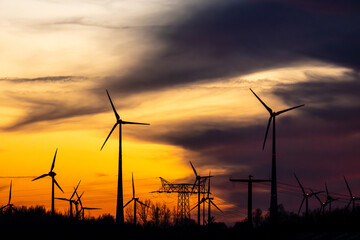 Obraz premium Wind turbines near the Leuna chemical works at sunset, silhouettes in a dramatic yellow and orange sky, with a high-voltage pylon and power lines emphasizing the energy landscape.