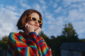 Teen girl (age 13) in a colorful sweater and yellow sunglasses, outdoor portrait under cloudy sky,...
