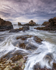 Obraz premium Long exposure seascape showing ocean water flowing over a rocky shoreline. Smooth water motion, coastal rocks, and cloudy sky create a natural marine landscape with dynamic movement.
