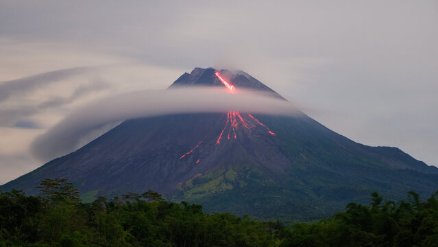 Red glowing lava flowing down the slopes of Mount Merapi while thick clouds swirl and cover the upper volcano. Dramatic volcanic activity creating a mysterious and powerful natural atmosphere.