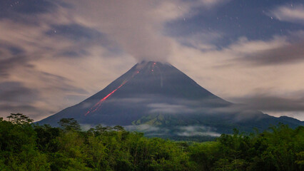 Fototapeta premium Active Volcano at Night With Glowing Lava Flow and Low Clouds