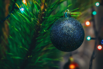 Shiny glitter Christmas ornament hanging on a tree with blurred needles and colorful lights in the background. Symbol of winter holidays, Christmas, New Year, celebration and festive mood.