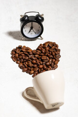 Coffee concept. A cup lies on a table with scattered coffee beans in a heart shape, a black alarm clock, a white background, top view