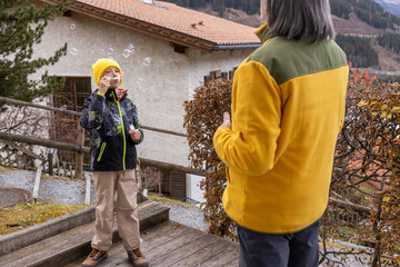 Child blows bubbles while standing on a wooden deck near a house in the mountains during daytime