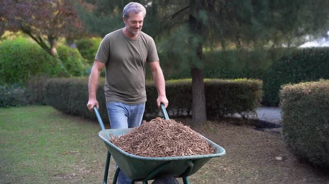 A man is pushing a wheelbarrow filled with wood chips in a lush garden, creating a scenic view